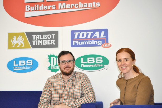 Ben and Rachel Davies sitting on blue sofas in front of a wall of LBS Group brand logos including Talbot Timber, Total Plumbing and LBS Renewables