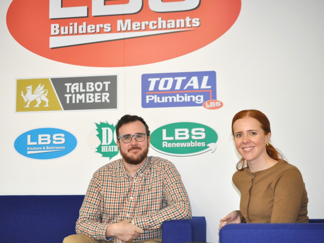Ben and Rachel Davies sitting on blue sofas in front of a wall of LBS Group brand logos including Talbot Timber, Total Plumbing and LBS Renewables