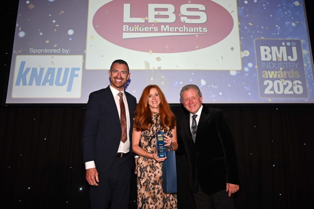Rachel Davies holding a blue BMJ Industry Awards 2026 trophy on stage between two men in suits, with the LBS Builders Merchants logo on the screen behind