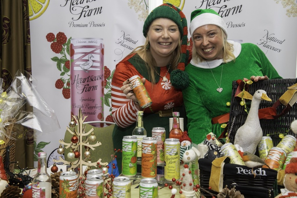 Two women in elf costumes smile behind a table of Heartsease Farm cans and bottles decorated for Christmas, with a Heartsease Farm banner behind them