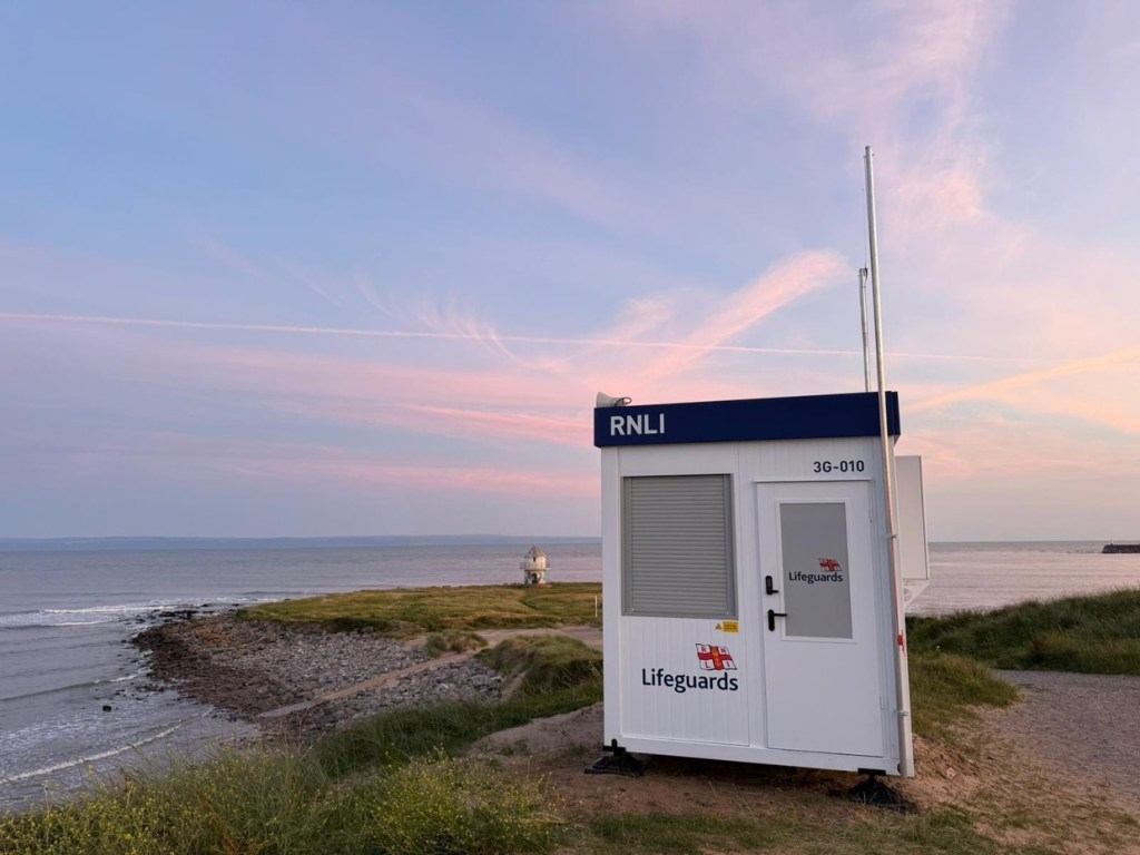 A white and blue RNLI beach lifeguard unit on a grassy shore at dusk, with the Porthcawl breakwater lighthouse behind
