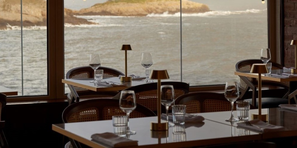 Tables set with wine glasses and brass lamps inside ROK restaurant, with Mumbles Head and the sea through the windows