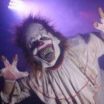 A scare actor in an evil clown mask and ragged white costume reaching towards the camera in purple stage lighting
