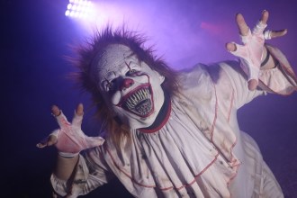 A scare actor in an evil clown mask and ragged white costume reaching towards the camera in purple stage lighting