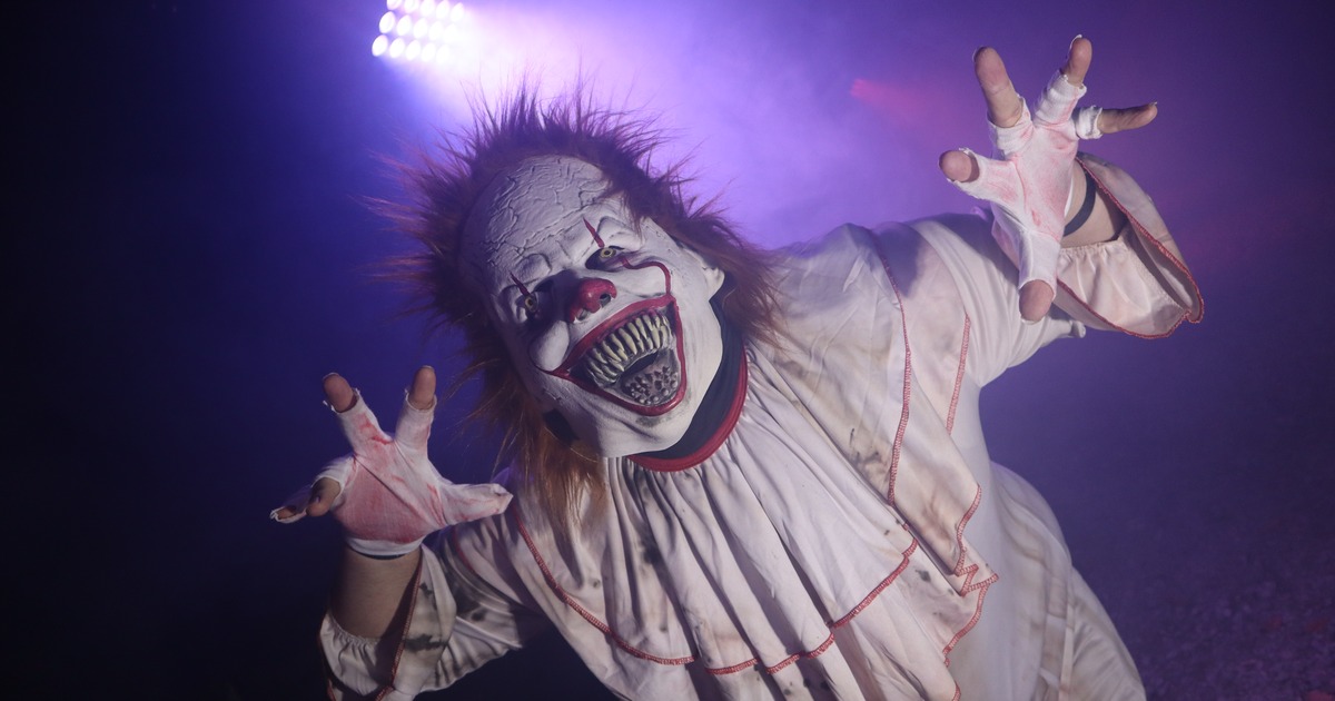 A scare actor in an evil clown mask and ragged white costume reaching towards the camera in purple stage lighting