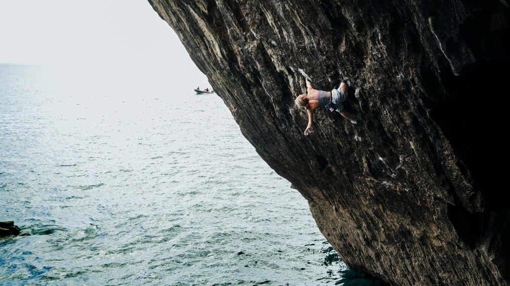 Shauna Coxsey on the overhanging cliff with a small support boat visible on the sea in the distance