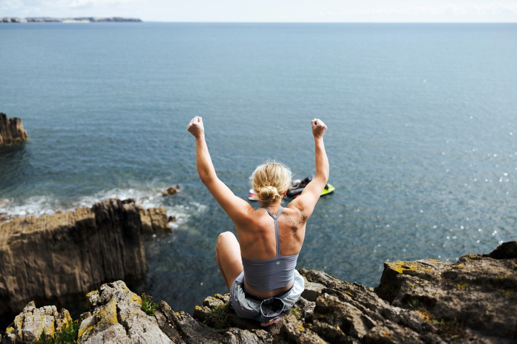 Shauna Coxsey sitting on the clifftop with both arms raised above her head, looking out to sea