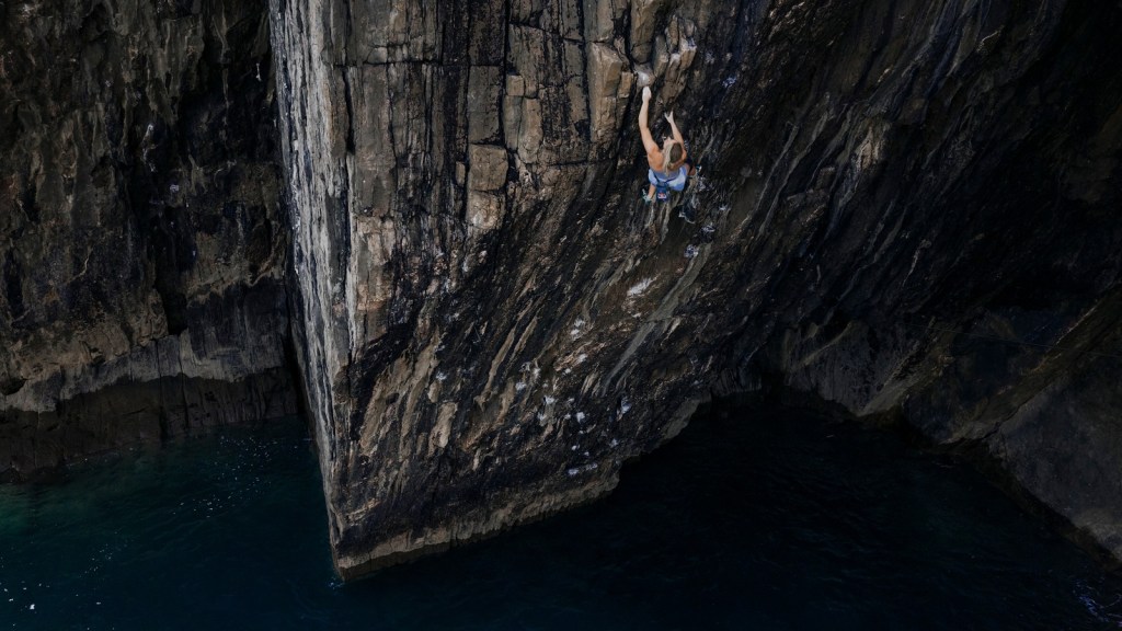 Aerial view of Shauna Coxsey part-way up the dark overhanging wall of The Olympiad with the sea beneath her