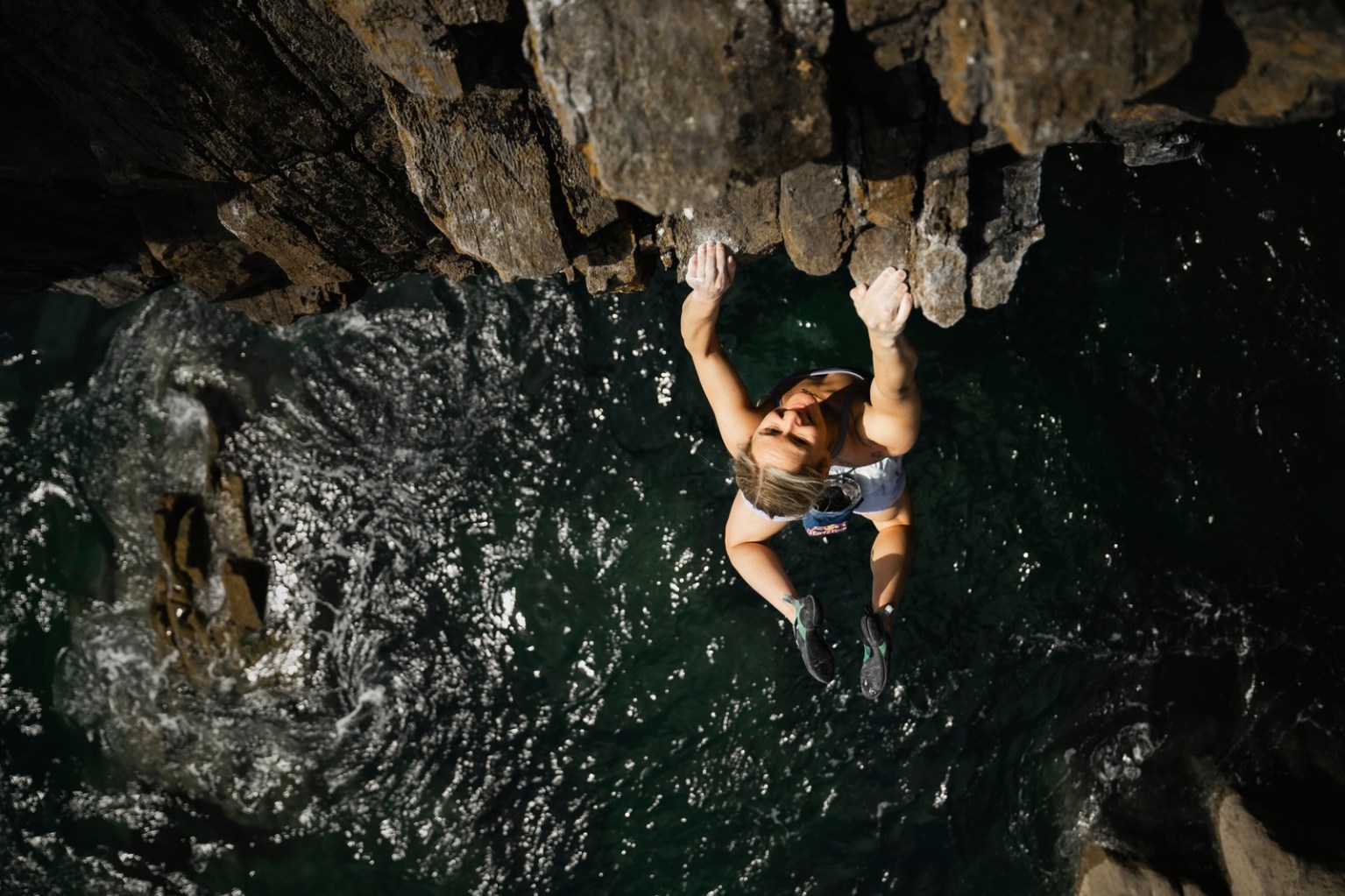 Shauna Coxsey hanging beneath the overhanging roof of The Olympiad at Skrinkle Haven, seen from above with the sea below her