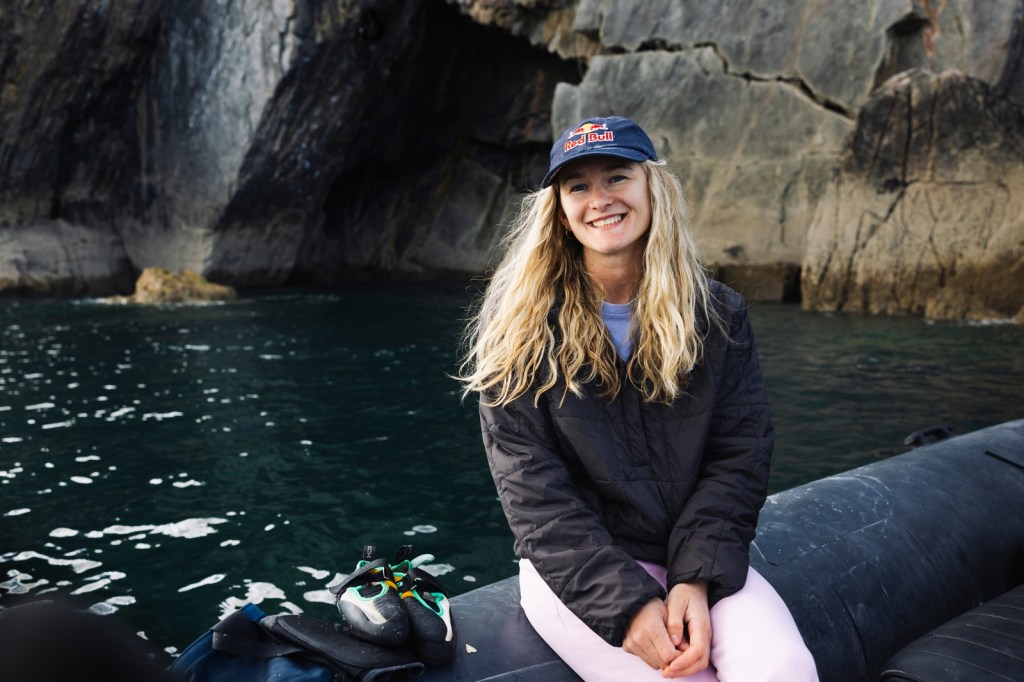 Shauna Coxsey sitting on the side of an inflatable boat beneath the cliffs, smiling, with her climbing shoes beside her