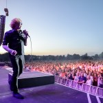 A singer with curly red hair and sunglasses holds a microphone at the edge of an outdoor stage at sunset, with a large crowd behind a barrier