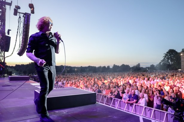 A singer with curly red hair and sunglasses holds a microphone at the edge of an outdoor stage at sunset, with a large crowd behind a barrier
