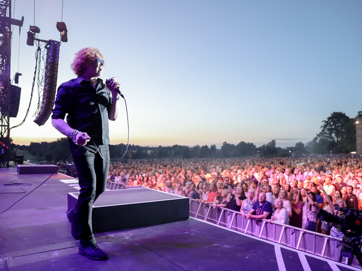 A singer with curly red hair and sunglasses holds a microphone at the edge of an outdoor stage at sunset, with a large crowd behind a barrier