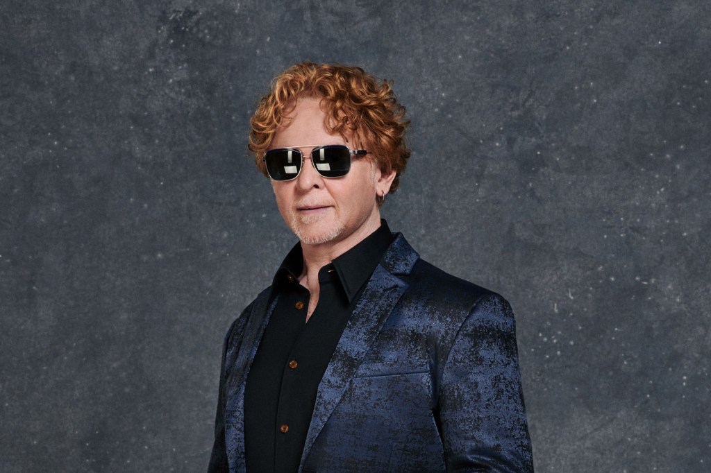 Studio portrait of a man with curly red hair and mirrored sunglasses, wearing a dark blue patterned jacket over a black shirt, against a grey backdrop
