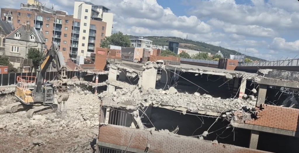 Rubble and exposed concrete decks of the part-demolished car park with a tower of flats and the hills of Swansea beyond