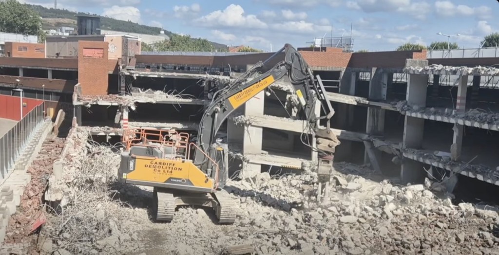 A demolition excavator with a breaker attachment working inside the stripped concrete frame of the car park