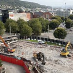 Aerial view of the flattened St David's car park site covered in crushed concrete, with excavators and workers in orange, the Vue cinema and tree-lined roads behind