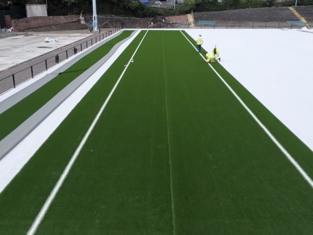Green artificial turf being rolled out in strips across the white underlay at St Helen's, with workers in hi-vis alongside