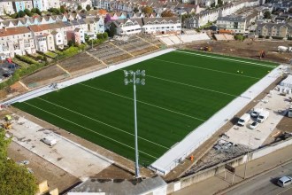 Aerial view of St Helen's in Swansea showing the new green artificial pitch marked out, a floodlight pylon in the foreground, the terrace and old stand on one side and construction work around the edges