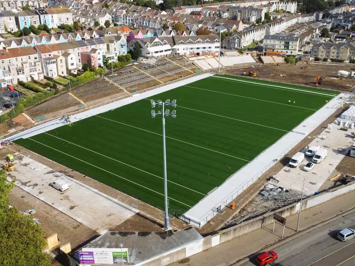 Aerial view of St Helen's in Swansea showing the new green artificial pitch marked out, a floodlight pylon in the foreground, the terrace and old stand on one side and construction work around the edges