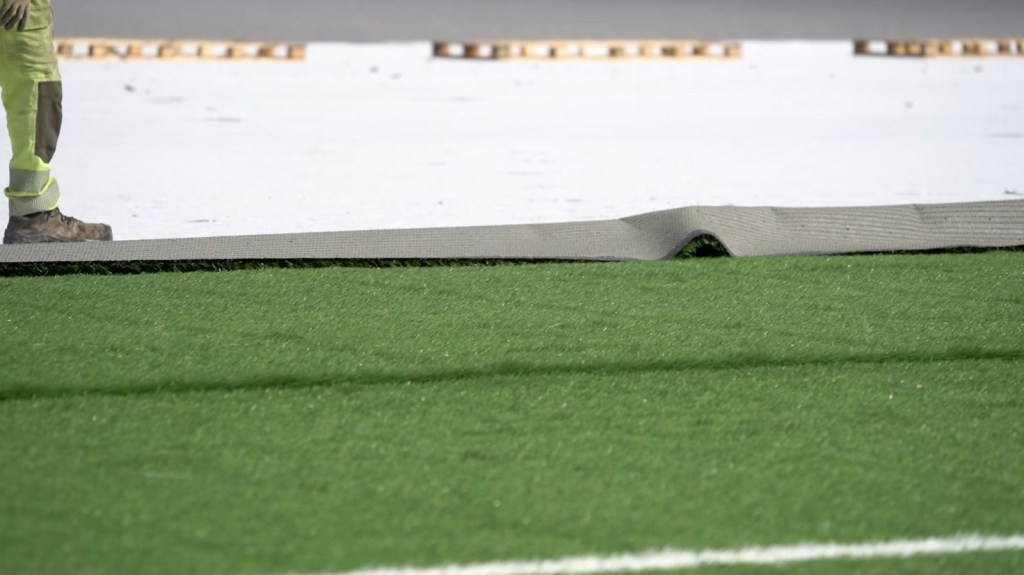 Close-up of a roll of green artificial turf being unrolled across white underlay, with a worker's boots beside it