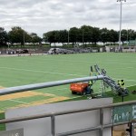 A galvanised floodlight column lying across the green artificial pitch at St Helen's with workers fitting lamp heads to it, an old lattice floodlight pylon standing in the background
