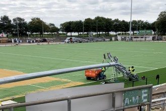 A galvanised floodlight column lying across the green artificial pitch at St Helen's with workers fitting lamp heads to it, an old lattice floodlight pylon standing in the background