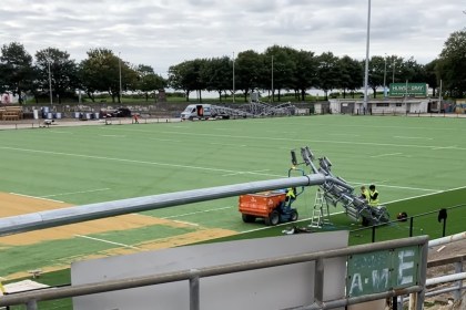 A galvanised floodlight column lying across the green artificial pitch at St Helen's with workers fitting lamp heads to it, an old lattice floodlight pylon standing in the background