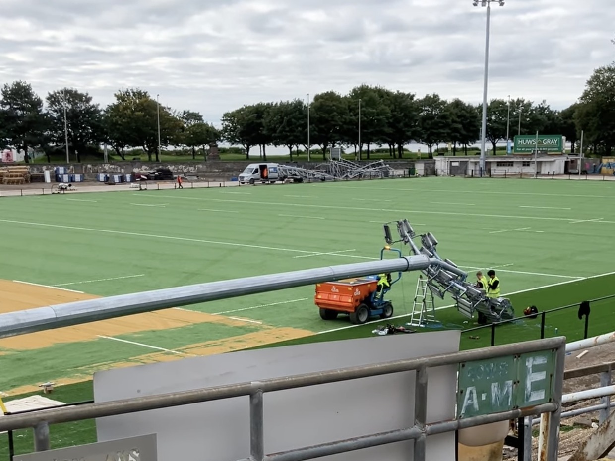 A galvanised floodlight column lying across the green artificial pitch at St Helen's with workers fitting lamp heads to it, an old lattice floodlight pylon standing in the background