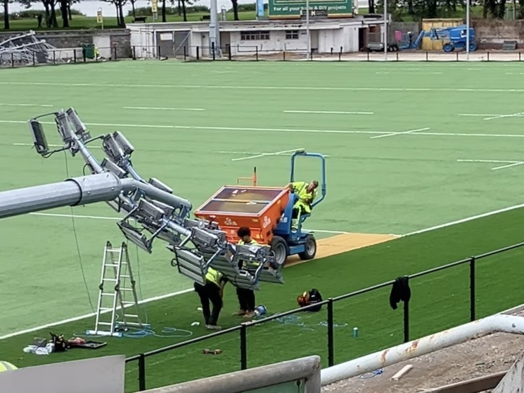 A worker drives an orange and blue sand spreader across the artificial pitch past a team fitting floodlight heads to a column on the ground