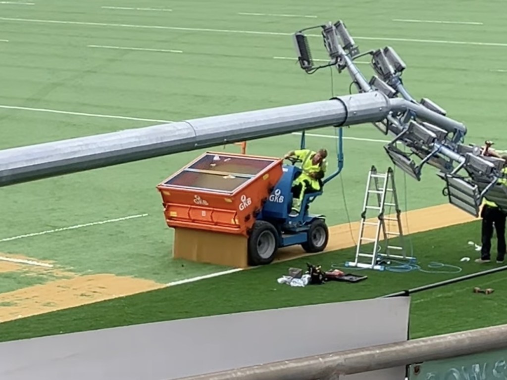 Close-up of workers in hi-vis fitting LED floodlight heads to the crossarm of a column lying on the pitch, with a ladder and an orange sand spreader beside them