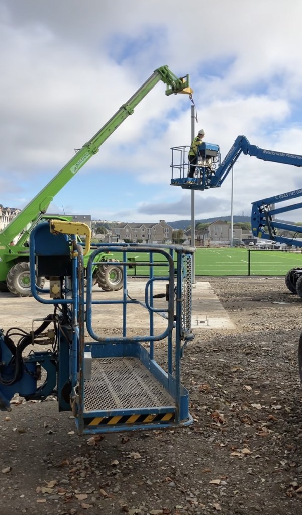 A green telehandler and a blue cherry picker raise a steel column onto a concrete pad beside the new pitch, with houses and hills behind