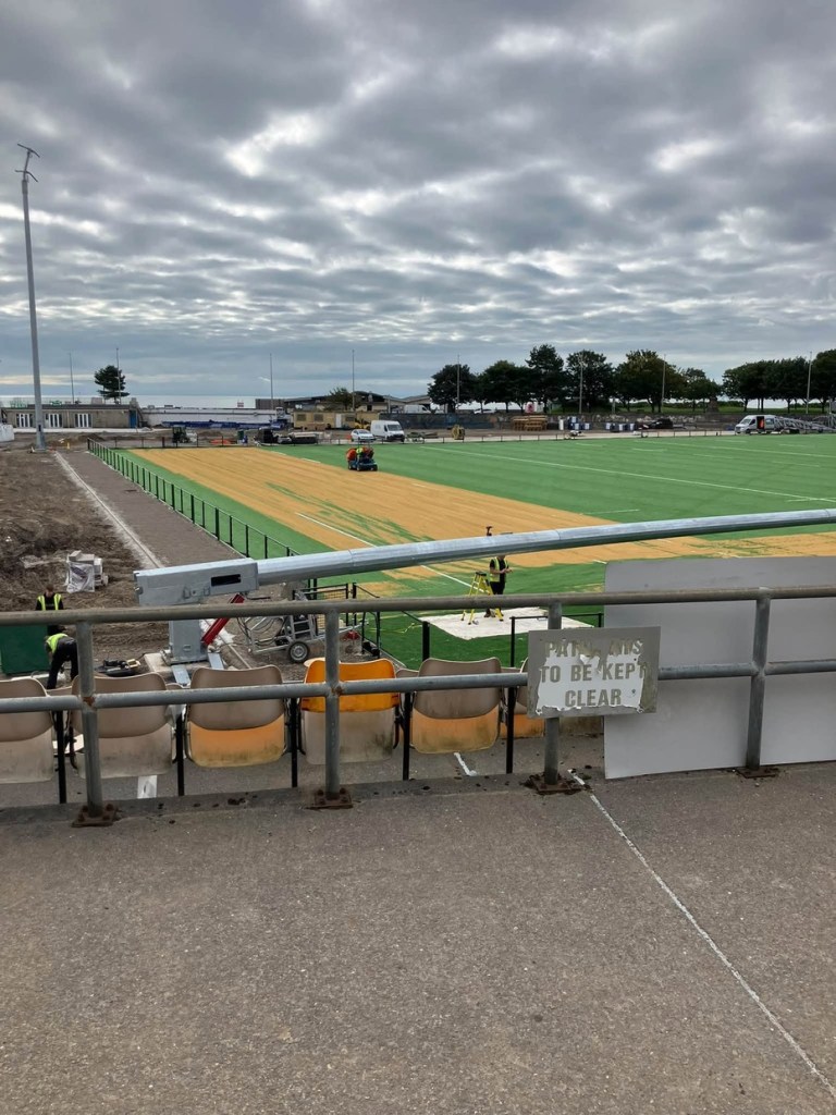 A row of faded orange and brown seats along a rail above the pitch, next to a peeling sign, with the sand-covered artificial pitch and grey sky beyond