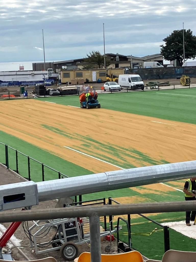 A sand spreader drives across the artificial pitch leaving a wide band of sand across the green surface, with site cabins and vans behind