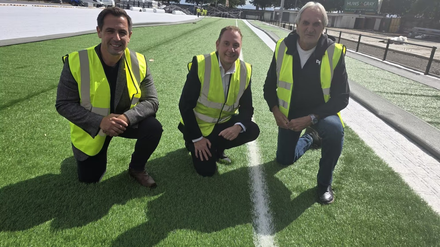 Three men in high-visibility vests kneeling on the new green artificial pitch at St Helen's, with the rest of the surface still being laid behind them