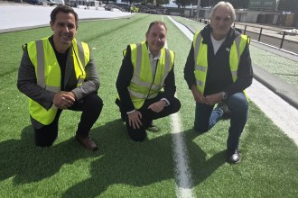Three men in high-visibility vests kneeling on the new green artificial pitch at St Helen's, with the rest of the surface still being laid behind them