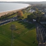 Aerial view of St Helen's rugby ground in Swansea before its redevelopment, with a grass pitch, a tall floodlight pylon, an uncovered terrace along the right-hand side and Swansea Bay behind