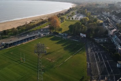 Aerial view of St Helen's rugby ground in Swansea before its redevelopment, with a grass pitch, a tall floodlight pylon, an uncovered terrace along the right-hand side and Swansea Bay behind