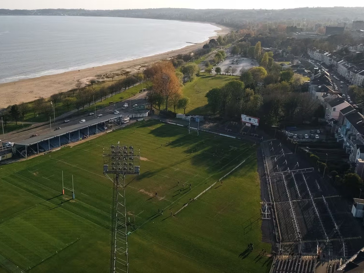 Aerial view of St Helen's rugby ground in Swansea before its redevelopment, with a grass pitch, a tall floodlight pylon, an uncovered terrace along the right-hand side and Swansea Bay behind