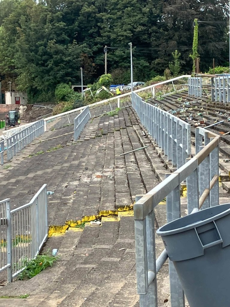 The old concrete terrace at St Helen's stripped of seats, with new galvanised barriers, weeds between the steps and a digger working at the far end