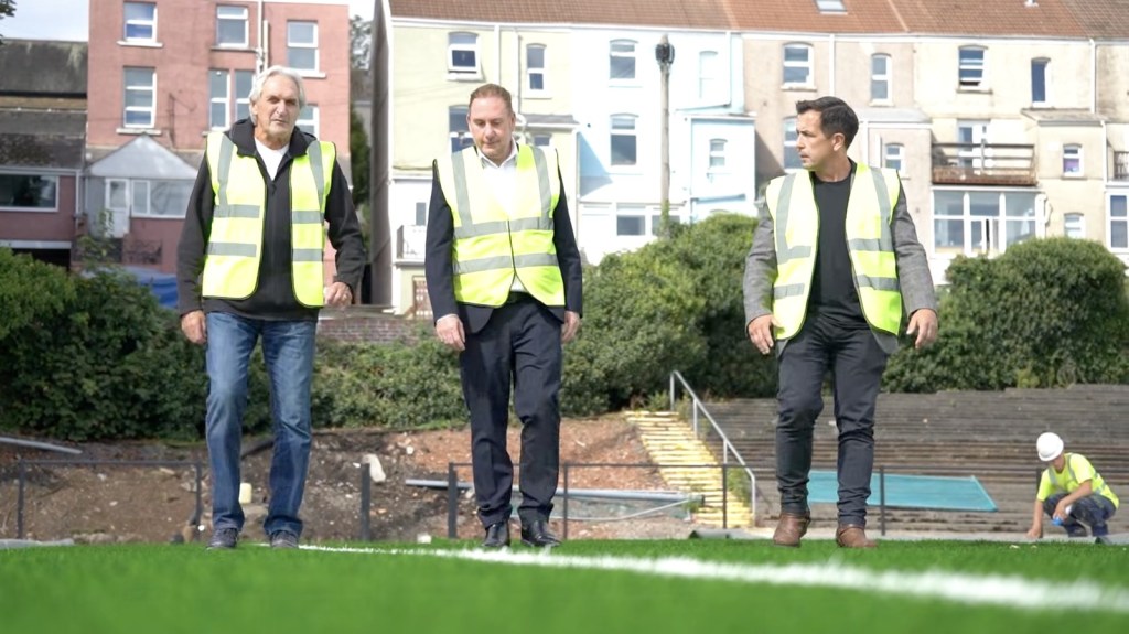 Three men in high-visibility vests walking across the new artificial pitch at St Helen's, with houses behind the ground