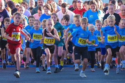 Dozens of children in blue race t-shirts and numbered bibs sprinting from the start line, with adults running behind