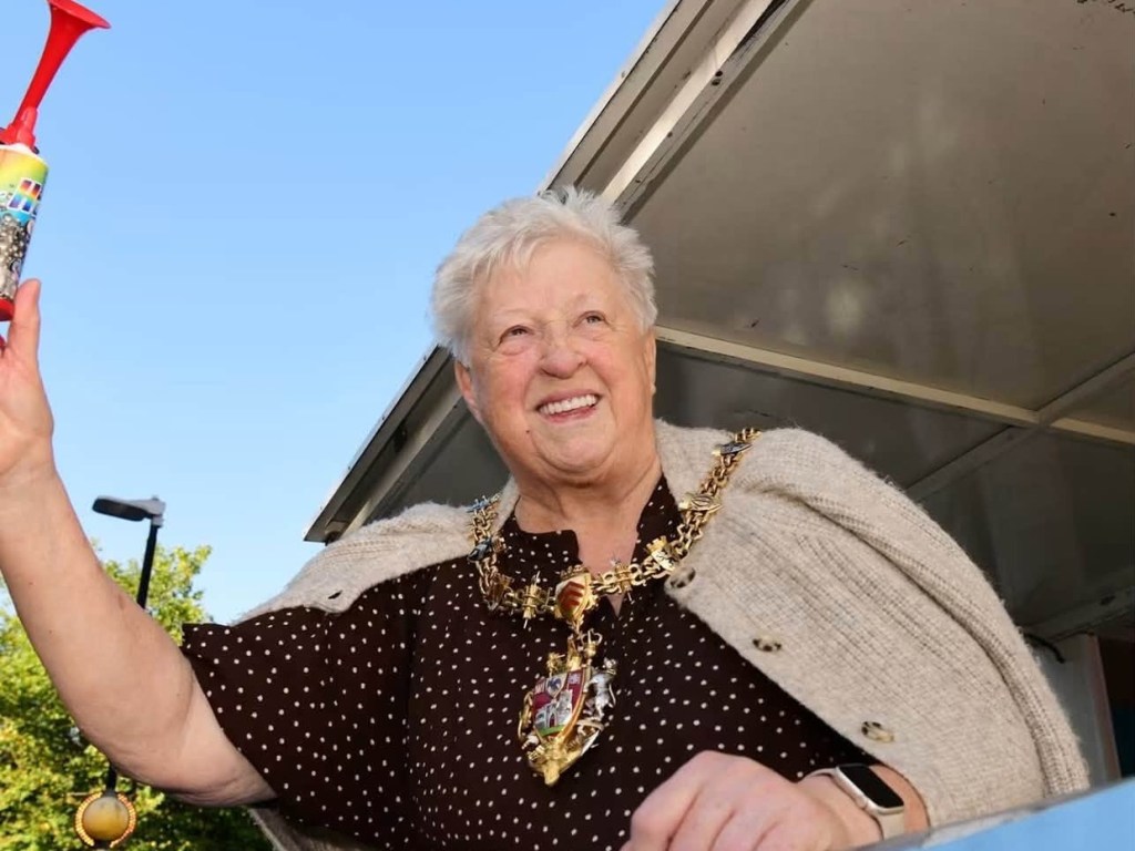 Smiling Lord Mayor Cllr Penny Matthews in her chain of office and a cream cardigan, holding an air horn aloft