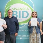 Six people holding BSL course certificates in front of a Swansea BID banner