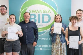Six people holding BSL course certificates in front of a Swansea BID banner