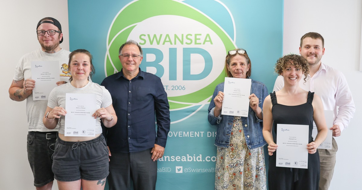 Six people holding BSL course certificates in front of a Swansea BID banner