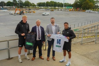 Two rugby players in Ospreys training kit holding sponsored shirts alongside two men in suits, standing above the stripped St Helen's pitch with construction plant behind