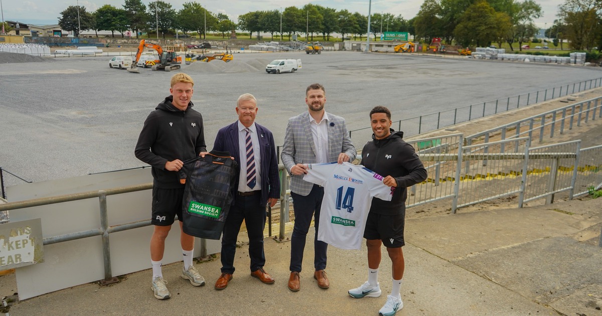 Two rugby players in Ospreys training kit holding sponsored shirts alongside two men in suits, standing above the stripped St Helen's pitch with construction plant behind