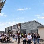 Artist's impression of supporters in Swansea City shirts outside a long grey-clad fan zone building with a club crest and player graphics, beside the stadium's white steel frame