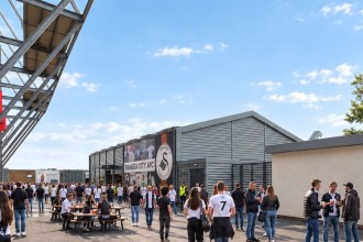 Artist's impression of supporters in Swansea City shirts outside a long grey-clad fan zone building with a club crest and player graphics, beside the stadium's white steel frame